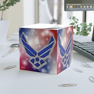 May include: A square memo cube featuring the United States Air Force logo in blue and silver on a red, white, and blue bokeh background. The emblem is displayed on two visible sides of the cube, which has a stack of white paper sheets on top.