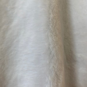 May include: White faux fur fabric with a soft, fluffy texture. The fur is long and dense, creating a luxurious look and feel.