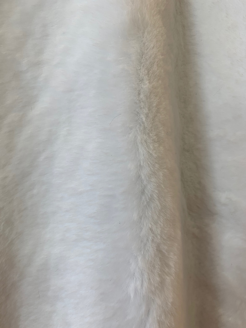 May include: White faux fur fabric with a soft, fluffy texture. The fur is long and dense, creating a luxurious look and feel.
