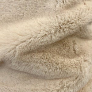 Può includere: Primo piano di un tessuto in finta pelliccia beige. Il tessuto ha una consistenza morbida e soffice con fibre lunghe e fluenti. Il colore è un beige chiaro e neutro, con sottili variazioni di tono, creando un senso di profondità e dimensione. La pelliccia è piegata.