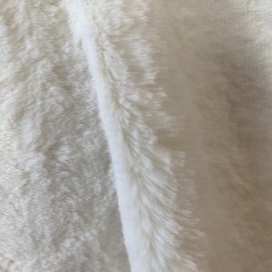 May include: A close-up of a white faux fur fabric. The fabric has a soft, fluffy texture and a subtle sheen.