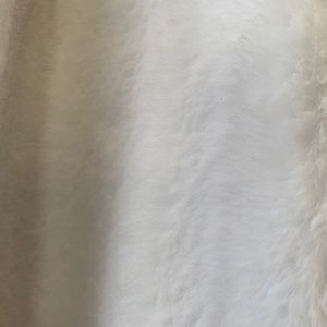 May include: White faux fur fabric with a soft, fluffy texture. The fur is arranged in a wavy pattern, creating a subtle, elegant look.