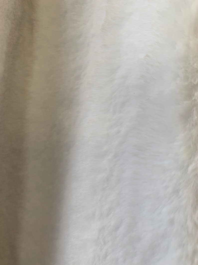 May include: White faux fur fabric with a soft, fluffy texture. The fur is arranged in a wavy pattern, creating a subtle, elegant look.