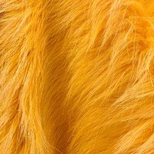 May include: Close-up of a textured, golden-yellow faux fur fabric. The fabric has long, soft fibers with subtle variations in color and texture. Thin, gold metallic threads are interspersed throughout, adding a touch of sparkle and shine. The overall effect is warm and inviting.