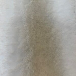 May include: A close-up of a white and beige faux fur fabric. The fur is soft and fluffy, with a gradient from white to beige.