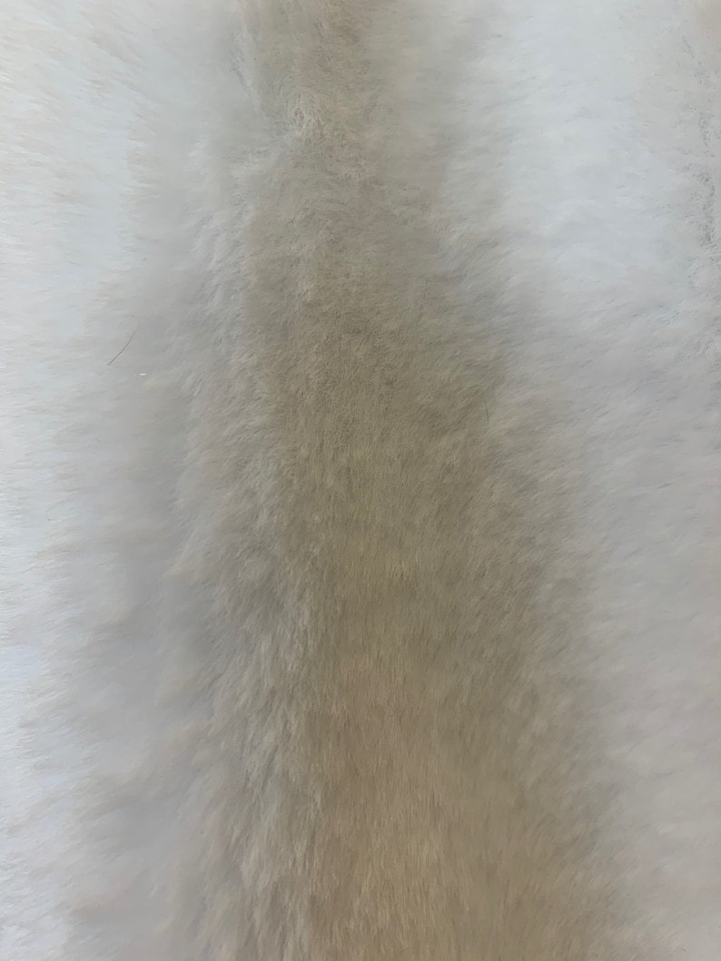 May include: A close-up of a white and beige faux fur fabric. The fur is soft and fluffy, with a gradient from white to beige.
