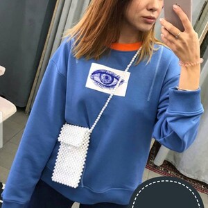 May include: A blue sweatshirt with an eye graphic and an orange trim around the neck. A white pearl phone case is worn as a crossbody bag. The text "PEARL PHONE CASE" is in a speech bubble.