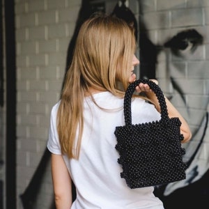 May include: A woman is holding a black beaded handbag. The bag has a square shape and is made of small, round beads.