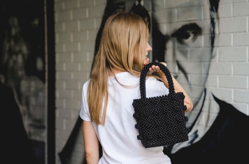 May include: A woman is holding a black beaded handbag. The bag has a square shape and is made of small, round beads.