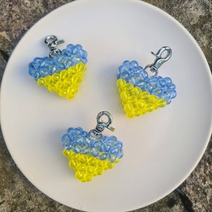 May include: Three heart-shaped keychains, each crafted from blue and yellow beads, arranged on a white plate. The keychains have silver-colored clasps. The color scheme mirrors the Ukrainian flag.