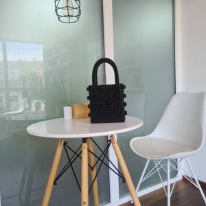May include: A black beaded handbag with a top handle sits on a white table with wooden legs. A white chair with black metal legs is in the background.