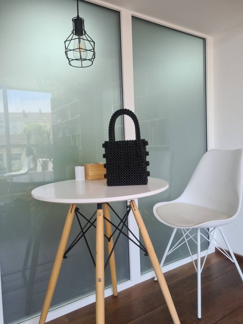 May include: A black beaded handbag with a top handle sits on a white table with wooden legs. A white chair with black metal legs is in the background.