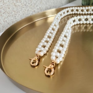 May include: A decorative bag strap constructed from interconnected white faux pearl beads, featuring polished gold-tone metal lobster claw clasps at both ends for secure attachment to a handbag or accessory.