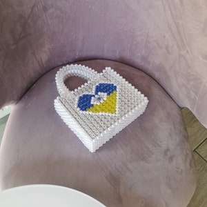 Puede incluir: Un bolso de mano de cuentas blancas con un diseño de corazón azul y amarillo. El bolso tiene un asa superior y está hecho de pequeñas cuentas redondas.