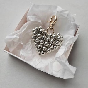 Peut inclure: Un porte-clés en forme de cœur argenté composé de petites perles rondes. Le porte-clés a un crochet doré.