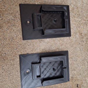 May include: Two black 3D printed wall mounts with a rectangular shape and a small hole in the center. The mounts have a raised lip on the top and bottom.