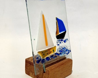 May include: A glass art piece depicting two sailboats on a blue and white background. The sailboats are made of stained glass and are mounted on a wooden stand.
