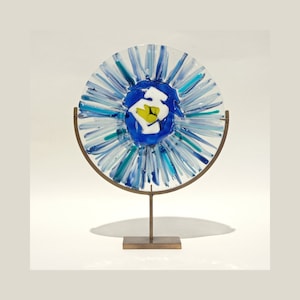 Op de afbeelding: Een blauw en wit glazen kunstwerk met een geel centrum, gemonteerd op een metalen standaard. Het glaswerk heeft een zonnestralenontwerp met blauwe en witte strepen.
