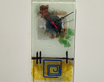 May include: A glass clock with a yellow, green, and brown abstract design. The clock face has black hands and a red second hand. The clock is framed with a black border and has a black and white design at the bottom.