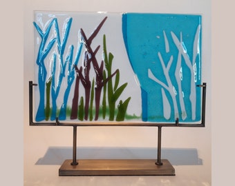May include: A glass art piece depicting a stylized landscape with blue, green, and white abstract trees on a white background. The piece is mounted on a metal stand with a wooden base.