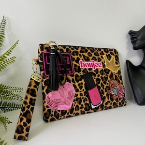 May include: A leopard print clutch bag with a wrist strap, featuring embroidered patches including a pink heart, a nail polish bottle, a gold crown, and text reading "boujee" and "FASHION JUNKIE." A black tassel charm hangs from the gold zipper pull.