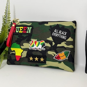 May include: Camouflage print clutch with multiple patches, including a green, yellow, and red map of Africa, a heart with the words "All Black Everything," and a red lip design. The clutch has a red wrist strap and the word "QUEEN" in green.