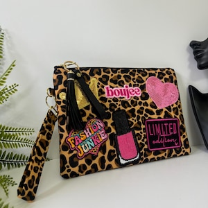 May include: A leopard print clutch bag with a wrist strap and black tassel. The bag features embroidered patches including a pink heart, a nail polish bottle, a gold crown, and text reading "boujee", "FASHION JUNKIE", and "LIMITED edition".