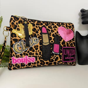 May include: A leopard print cosmetic bag with gold zipper and wrist strap. Embellished with sequined crown, lipstick, nail polish, and a pink heart. Text includes "Chic", "boujee", and "Limited Edition".