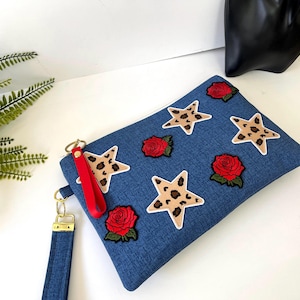 May include: A denim clutch with a zipper closure, adorned with leopard print star patches and red rose appliques. It features a red wrist strap and a gold-colored wristlet strap. The clutch is approximately 10 inches long and 6 inches high.