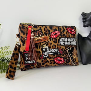 May include: A leopard print clutch bag featuring various patches including red lips, the text "HATE ME OR LOVE ME ONLY GOD CAN JUDGE ME", "SORRY NOT SORRY", and "Queen". It includes a brown tassel, a red strap, and a leopard print wristlet strap.