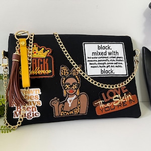 May include: Black clutch with gold chain strap and tassel. Features patches with text and illustrations, including "Black Excellence," a woman in sunglasses, and "Love The Skin You're In."