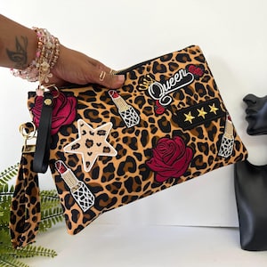 May include: A leopard print clutch with a black zipper, adorned with embroidered patches of a star, lipstick, roses, and the word "Queen". The clutch has a black wrist strap and gold-colored hardware.