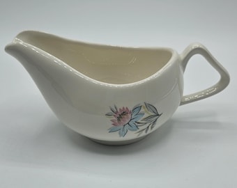 Puede incluir: Una salsera de cerámica de color crema con un pico vertedor puntiagudo y un asa angular. El exterior está decorado con un motivo floral que consiste en una flor rosa y azul claro con hojas grises situada en el lateral del recipiente.