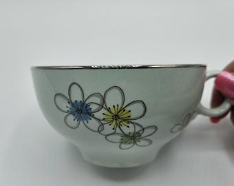 May include: A light green teacup with a silver rim and a handle. The cup is decorated with a floral design featuring blue, yellow, and white flowers. The teacup is likely made of ceramic or porcelain.