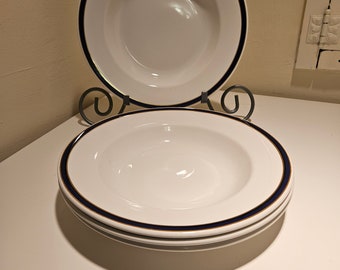 May include: A stack of white ceramic soup bowls with a navy blue and gold rim. The bowls are stacked on a white surface, with one bowl displayed on a decorative metal stand. The bowls have a deep center.