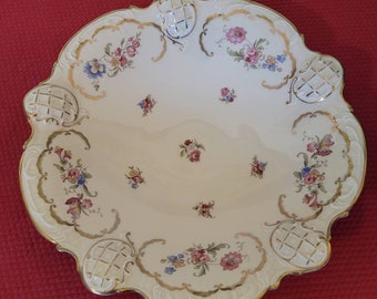 May include: A white porcelain serving bowl with a gold rim and floral design. The bowl has a scalloped edge and four decorative handles.
