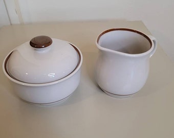 May include: A white ceramic sugar bowl with a lid and a small creamer. Both pieces have a brown trim and a brown knob on the lid. The set is on a light beige surface.