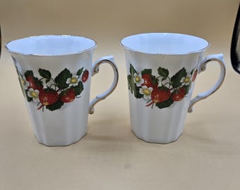 May include: Two white ceramic mugs with a strawberry design. Each mug features a gold rim and handle, and a pattern of red strawberries, green leaves, and white flowers. The mugs have a faceted shape.