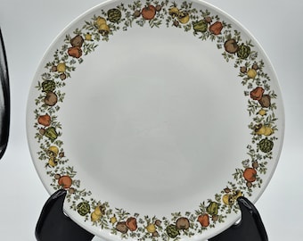 May include: White ceramic plate with a decorative border of colorful fruits and vegetables, including tomatoes, squash, and artichokes. The plate is displayed on a black stand against a white background. A vintage-style dinnerware.