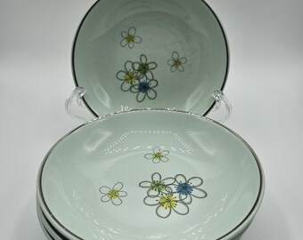 May include: A stack of four light green ceramic bowls with a floral design. Each bowl has a black rim and a silver-colored edge. The floral design features yellow, blue, and white flowers.