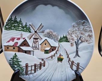 May include: A decorative ceramic plate featuring a winter village scene. The plate depicts snow-covered houses, a windmill, and a road with a cart. The color palette includes white, brown, green, and gray, creating a serene winter landscape.