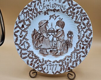 Puede incluir: Un plato decorativo con un diseño marrón y blanco, con las palabras "Merry Christmas" y el año "1977". El plato representa a dos niños sosteniendo un pastel, rodeados de motivos de acebo y bastones de caramelo, sobre un soporte de metal.