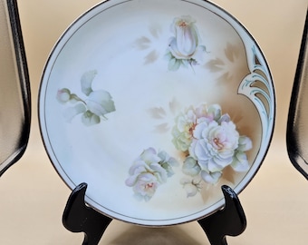 May include: A round, decorative plate with a floral design. The plate features white and pink roses with green leaves on a cream-colored background. The plate has a brown border and is displayed on a black stand.