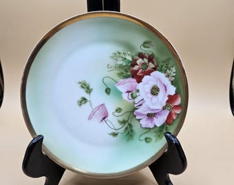 May include: A decorative porcelain plate with a gold rim, featuring hand-painted pink and red poppies on a pale green background. The plate is displayed on a black stand, showcasing the floral design and elegant details.