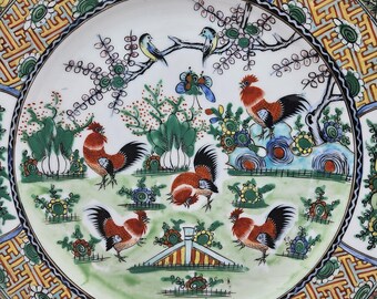 Puede incluir: Un plato de cerámica decorativo con un diseño detallado que presenta gallos, pájaros y elementos florales. El plato tiene una base blanca con un borde colorido y patrones intrincados en tonos de rojo, verde, amarillo y azul. El plato está sostenido por un soporte de metal.