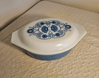 May include: A vintage casserole dish with a white lid and blue floral design. The dish has a light blue base and is oval-shaped with handles. The lid features a symmetrical pattern of stylized flowers and leaves.