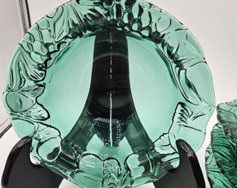 May include: A set of two teal-colored glass dishes with a floral design around the rim. The dishes are displayed on black stands, showcasing their intricate details and elegant shape. The glass has a glossy finish.