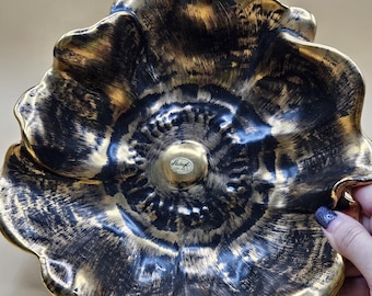 Puede incluir: Un cuenco decorativo de cerámica negra y dorada con forma de flor. El cuenco tiene una superficie texturizada con un borde y detalles dorados metálicos. El cuenco está sostenido por una mano con una uña pintada.