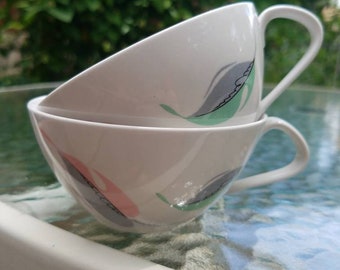 May include: Two white teacups stacked on top of each other. Each cup has a curved handle and is decorated with a leaf design in gray, pink, and green. The cups are on a glass table with a green leafy background.