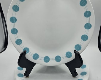 Puede incluir: Una pila de tres platos de cerámica blanca con un borde decorativo de círculos azul cerceta conectados por finas líneas discontinuas. Los platos son circulares, tienen un acabado liso y brillante, y se exhiben sobre un soporte de plástico negro.